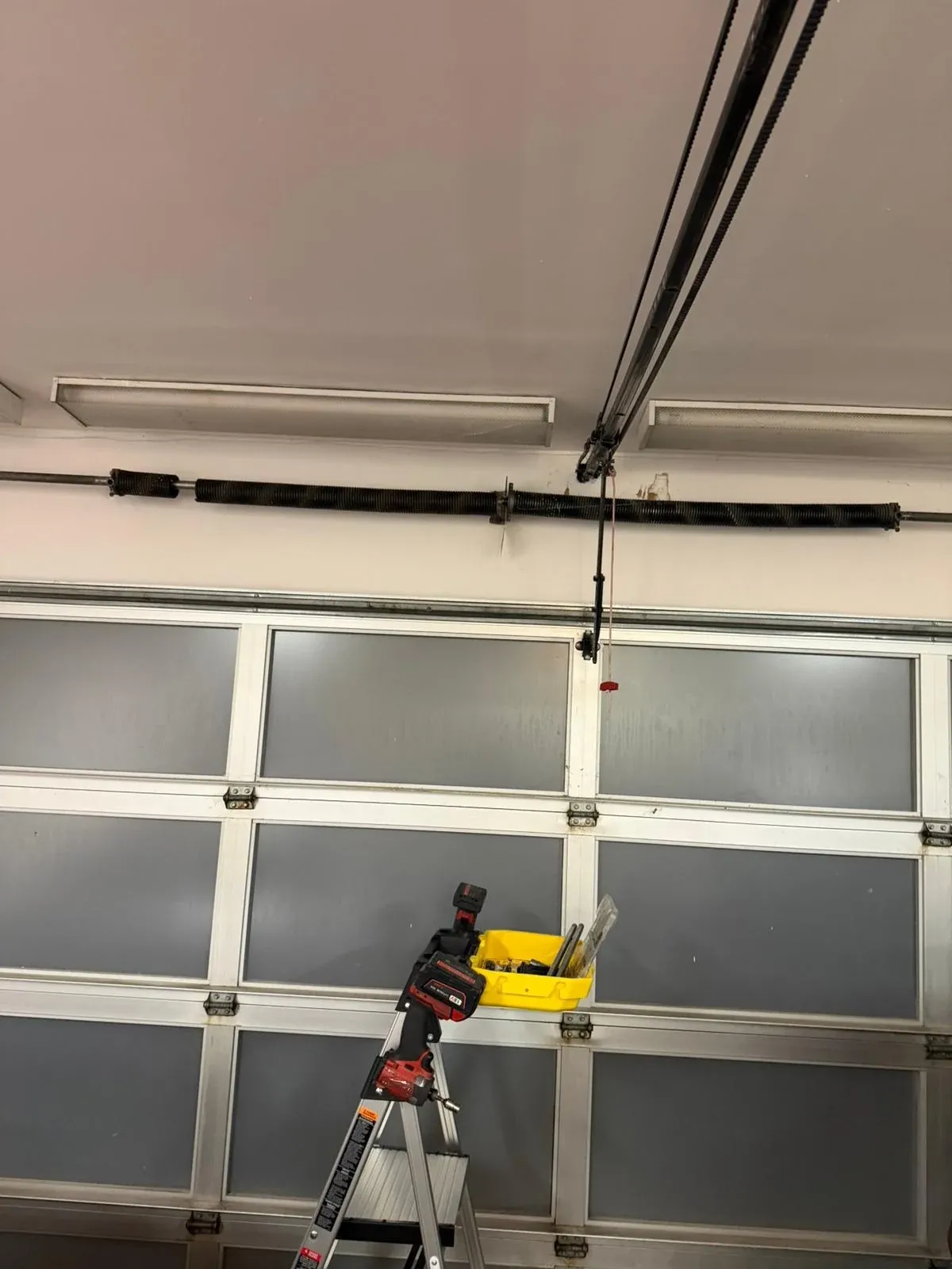 A full-view garage door with frosted glass panels, the torsion spring on the header above it and a stepladder with a yellow tool tray