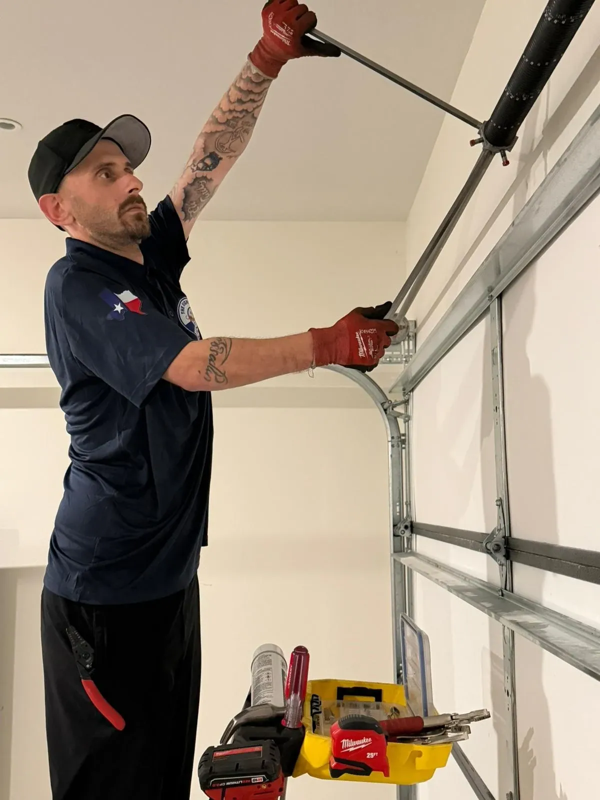 A technician in a navy company shirt with a Texas flag patch, on a ladder, reaching up with a winding bar in the torsion spring cone