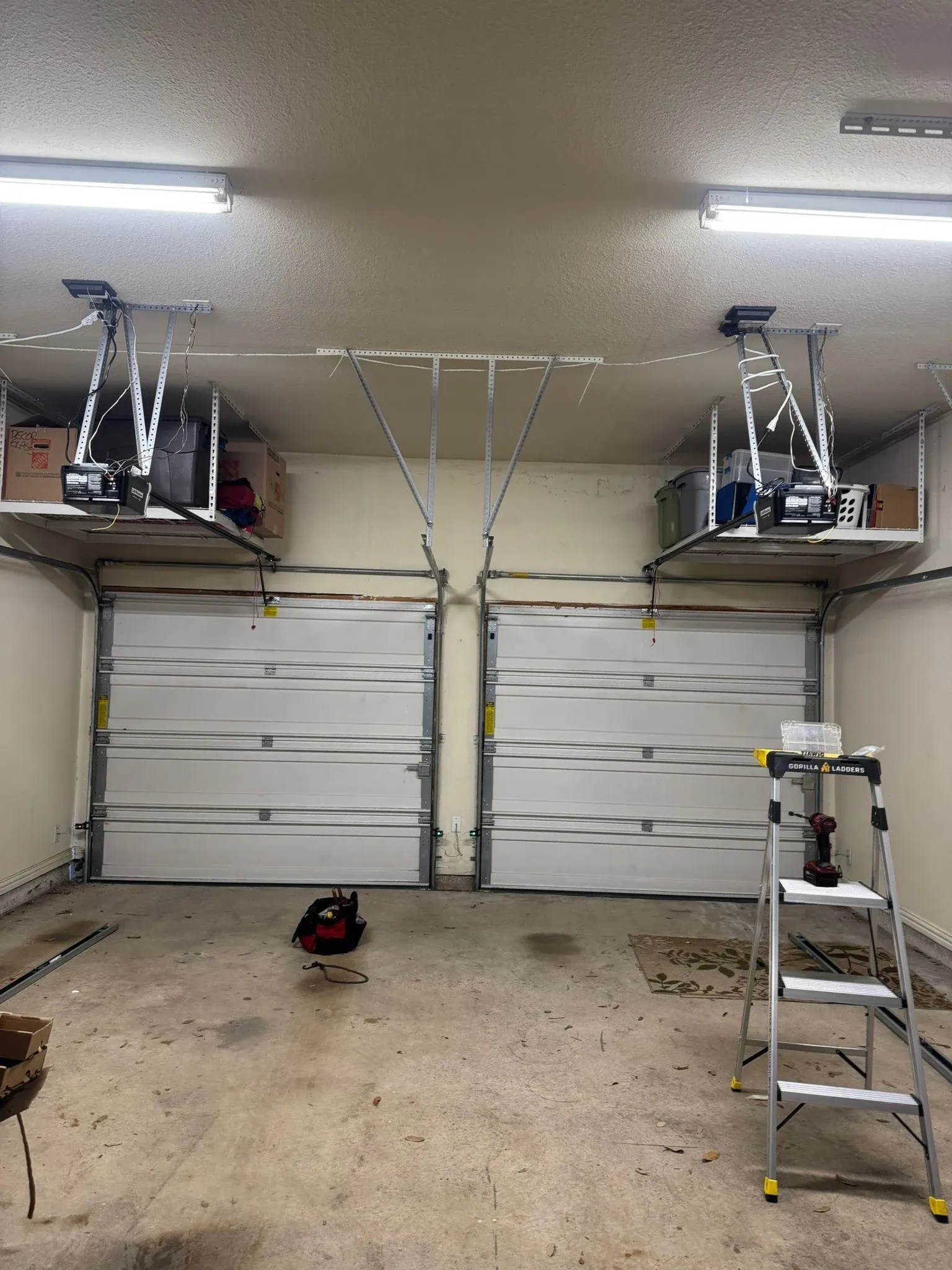 Two white single garage doors side by side, each with its own ceiling-mounted opener below overhead storage racks